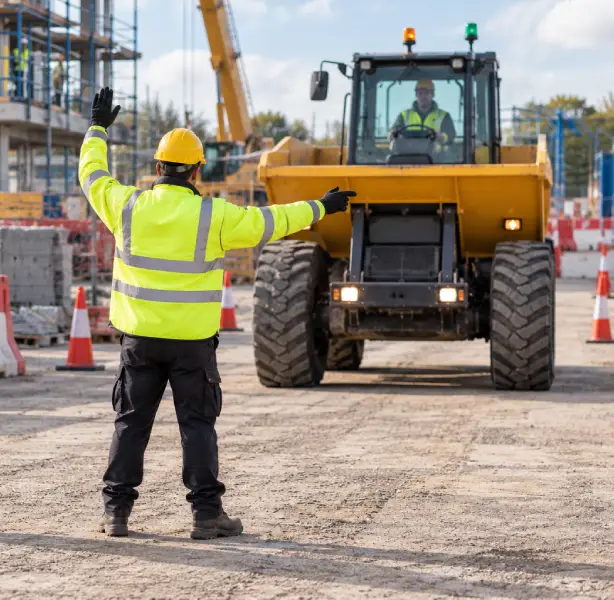 Traffic marshal in a hard hat and high visibility jacket signalling a dumper truck to stop on a construction site